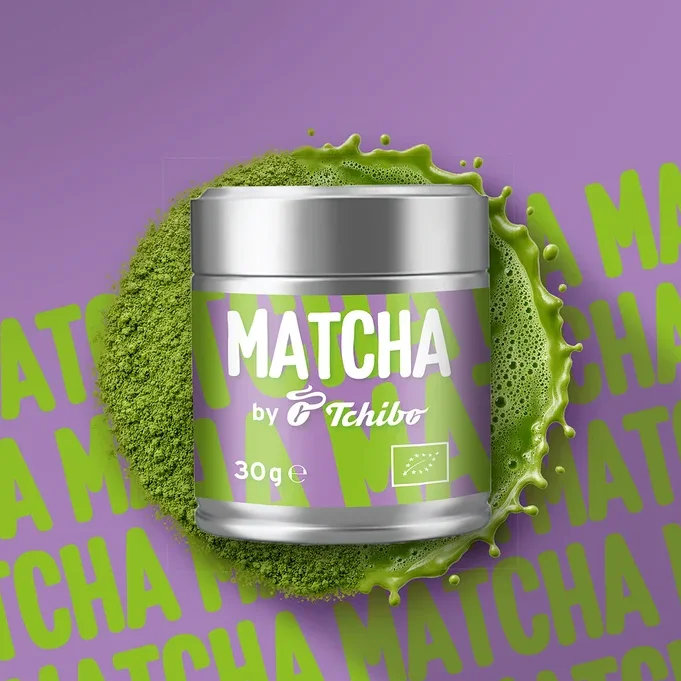 Silberne Dose Matcha von Tchibo mit lila Etikett, umgeben von grünem Matchapulver und Spritzern, auf lila Hintergrund mit wiederholtem Matcha-Schriftzug.