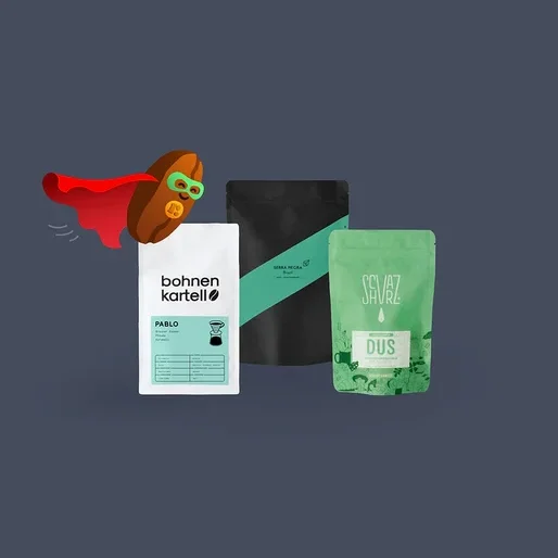Eine Comic-Kaffeebohne mit rotem Umhang und grüner Maske fliegt an drei Kaffeepackungen vorbei: einer weißen 'bohnen kartell'-Packung, einer schwarzen 'SIERRA NEGRA'-Packung und einer grünen 'SEVAZ DUS'-Packung.