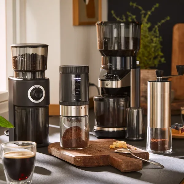 Vier Kaffeemühlen und eine Packung Kaffeebohnen sind auf einer Küchentheke mit Kaffee und Gebäck arrangiert.