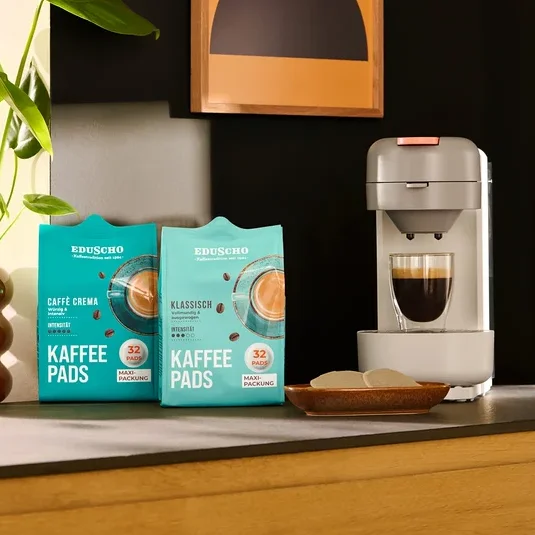 Grüne Pflanze in braunem Topf, zwei Packungen Eduscho Caffe Crema und Klassisch Kaffeepads, und eine hellgraue Kaffeemaschine mit rosa Knopf, die Kaffee in ein Glas füllt. Mehrere weiße Kaffeepads liegen in einer kleinen Holzschale, alles auf einer Küchenzeile.