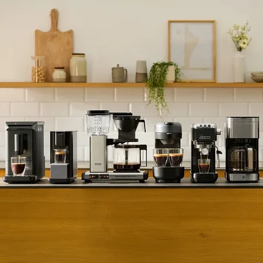Sechs verschiedene Kaffeemaschinen stehen auf einer dunklen Kücheninsel-Arbeitsplatte. Von links nach rechts sind zwei schwarze Einzelportionsmaschinen, eine silber-schwarze Filterkaffeemaschine, eine dunkelgraue Maschine mit zwei Tassen, eine schwarze Espressomaschine und eine weitere silber-schwarze Filterkaffeemaschine zu sehen. Einige Maschinen haben Tassen mit Kaffee darunter. Im Hintergrund befindet sich ein Holzregal mit Küchendekoration.