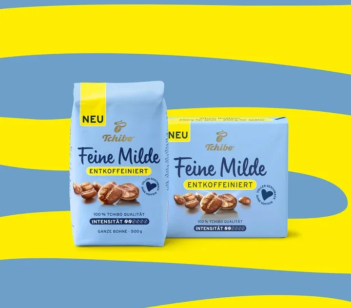 Zwei hellblaue Tchibo Feine Milde entkoffeinierte Kaffeepackungen, eine stehende 500g Bohnenpackung und eine liegende Packung, auf blau-gelbem Hintergrund.
