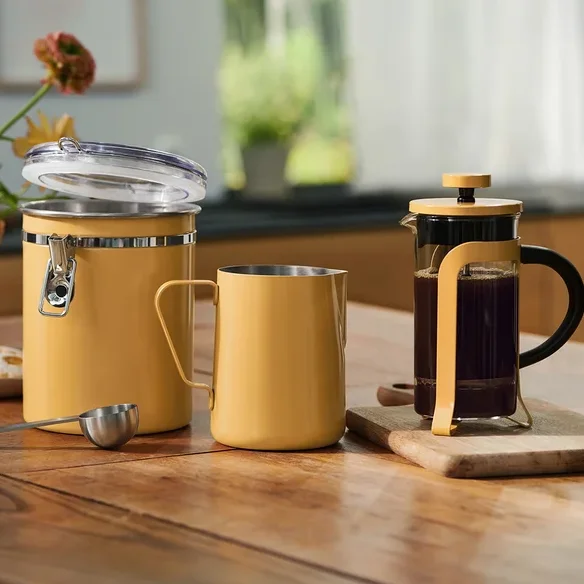 Ein gelber Behälter mit offenem Deckel, ein gelber Metallbecher und ein silberner Messlöffel stehen auf einem Holztisch. Rechts davon steht eine French Press mit Kaffee auf einem kleinen Schneidebrett. Eine Blumenvase und Kekse auf einem Teller befinden sich links.