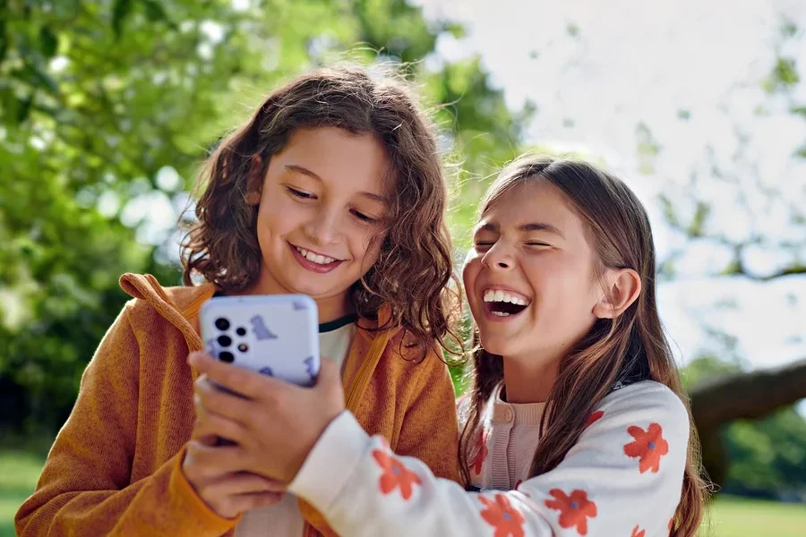 Zwei lachende Kinder schauen sich draußen ein Handy mit einer Dinosaurierhülle an.