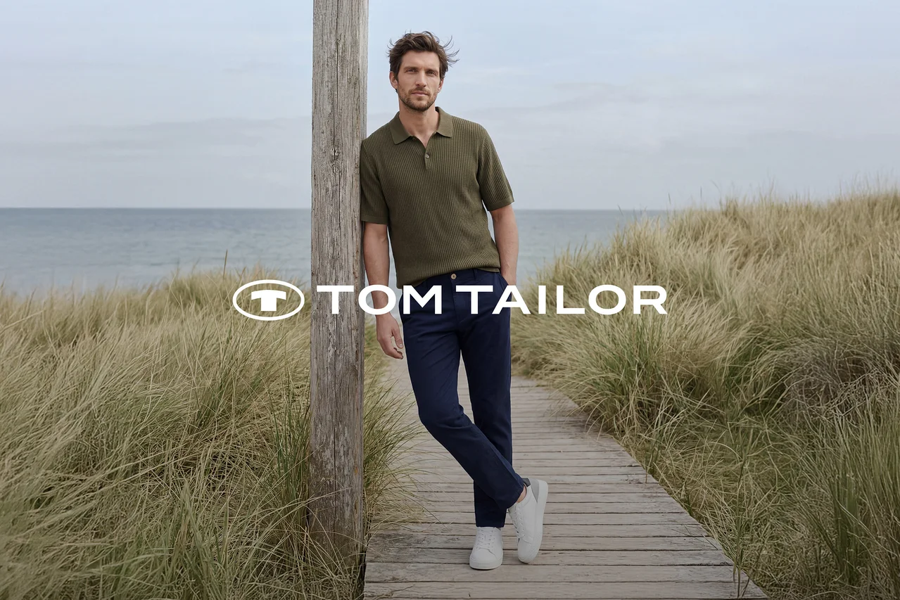 Mann in grünem Poloshirt, dunkelblauer Hose und weißen Sneakern lehnt an einem Holzpfahl auf einem Holzsteg am Meer, umgeben von hohem Gras. TOM TAILOR Logo.