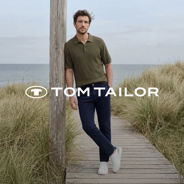 Mann in grünem Poloshirt, dunkelblauer Hose und weißen Sneakern lehnt an einem Holzpfahl auf einem Holzsteg am Meer, umgeben von hohem Gras. TOM TAILOR Logo.