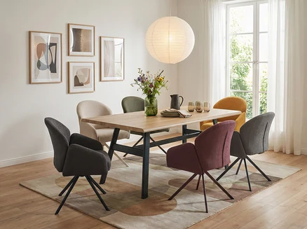 Ein Esszimmer mit einem Holztisch und sechs Stühlen in verschiedenen Farben auf einem gemusterten Teppich. Auf dem Tisch stehen eine Blumenvase, ein Krug und Geschirr. Eine große Papierlampe hängt über dem Tisch, und an der Wand hängen vier gerahmte Bilder.