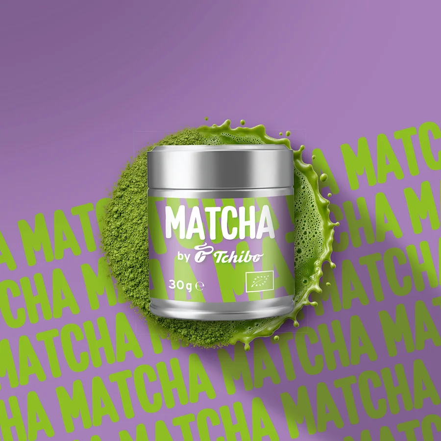 Silberne Dose Matcha von Tchibo mit lila Etikett, umgeben von grünem Matchapulver und Spritzern, auf lila Hintergrund mit wiederholtem Matcha-Schriftzug.