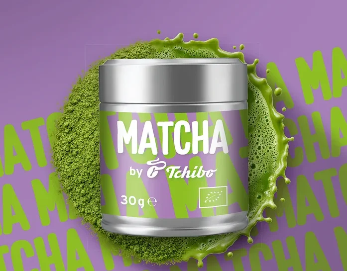 Silberne Dose Matcha von Tchibo mit lila Etikett, umgeben von grünem Matchapulver und Spritzern, auf lila Hintergrund mit wiederholtem Matcha-Schriftzug.