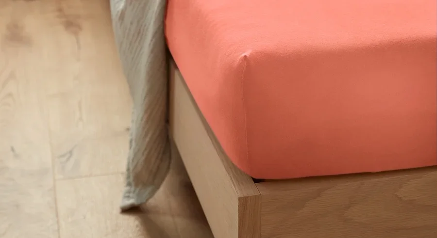Korallefarbenes Jersey-Spannbettlaken in Normalgröße auf einem Bett mit weißen Kissen und Überwurf.