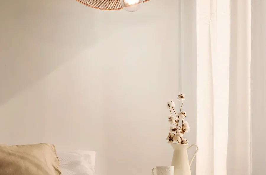 Eine strohfarbene Lampen-Hängelampe in Hutform hängt über einem Bett in einem Schlafzimmer.