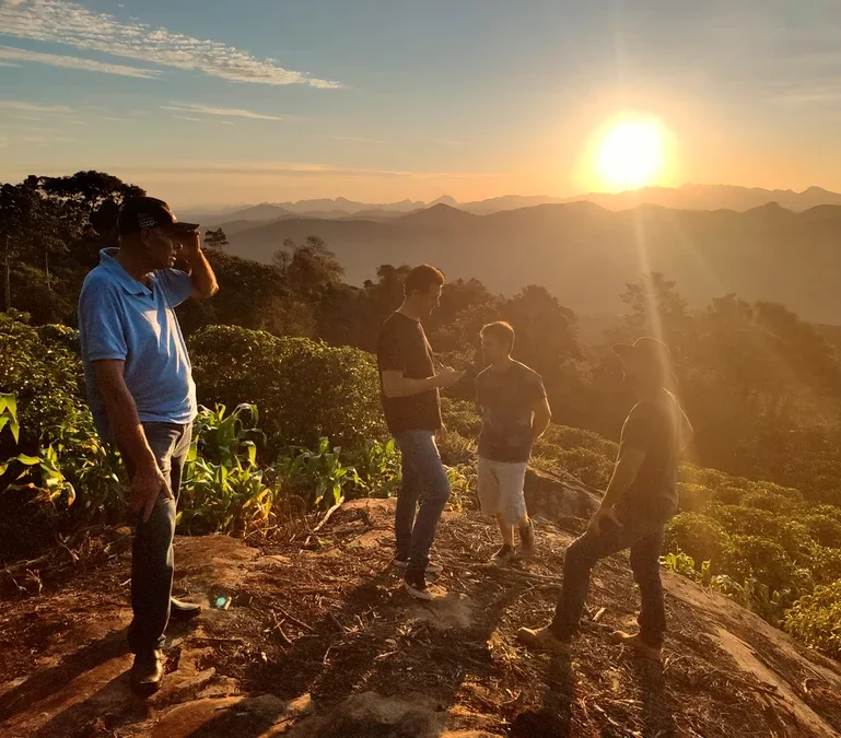 Vier Männer stehen auf einem Hügel bei Sonnenaufgang. Im Hintergrund sind Berge zu sehen.