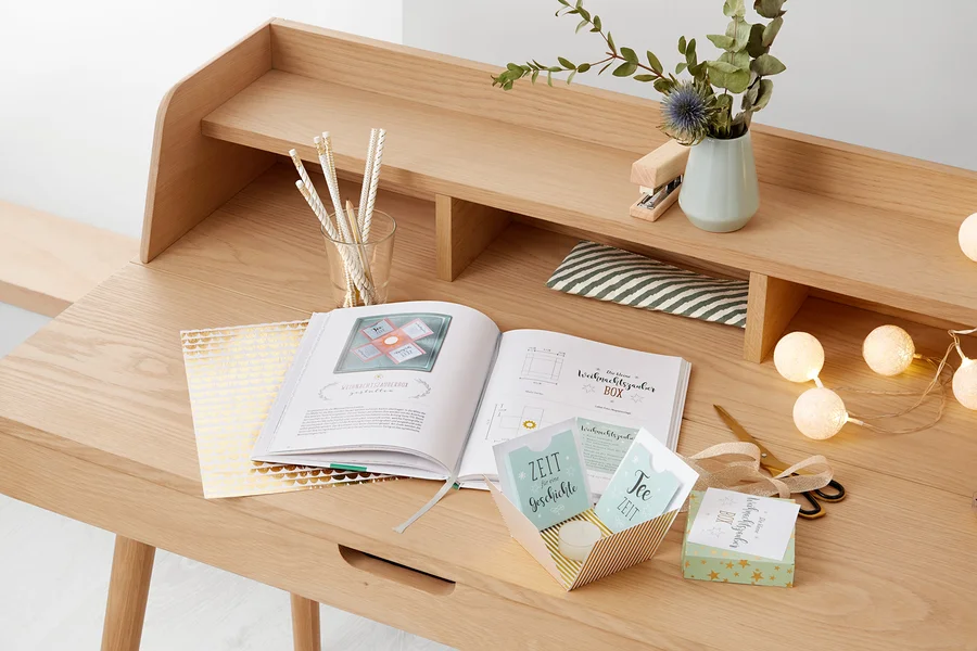 Ein aufgeschlagenes Buch mit einer Anleitung zur Herstellung einer Weihnachtsbox liegt auf einem Holztisch. Daneben stehen eine Vase mit Pflanzen und ein Glas mit Stiften.
