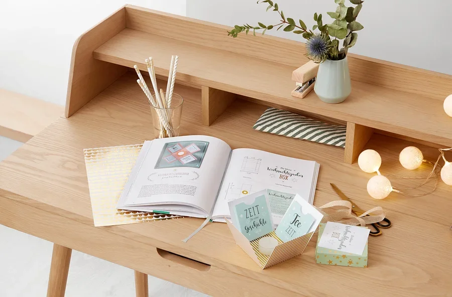 Ein aufgeschlagenes Buch mit einer Anleitung zur Herstellung einer Weihnachtsbox liegt auf einem Holztisch. Daneben stehen eine Vase mit Pflanzen und ein Glas mit Stiften.