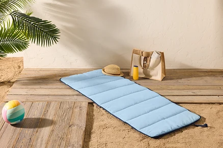 Eine hellblaue Strandmatte liegt auf einem Holzboden neben Sand, mit einem Strohhut, einer Sonnencremeflasche und einer Tragetasche in der Nähe.