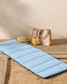 Eine hellblaue Strandmatte liegt auf einem Holzboden neben Sand, mit einem Strohhut, einer Sonnencremeflasche und einer Tragetasche in der Nähe.