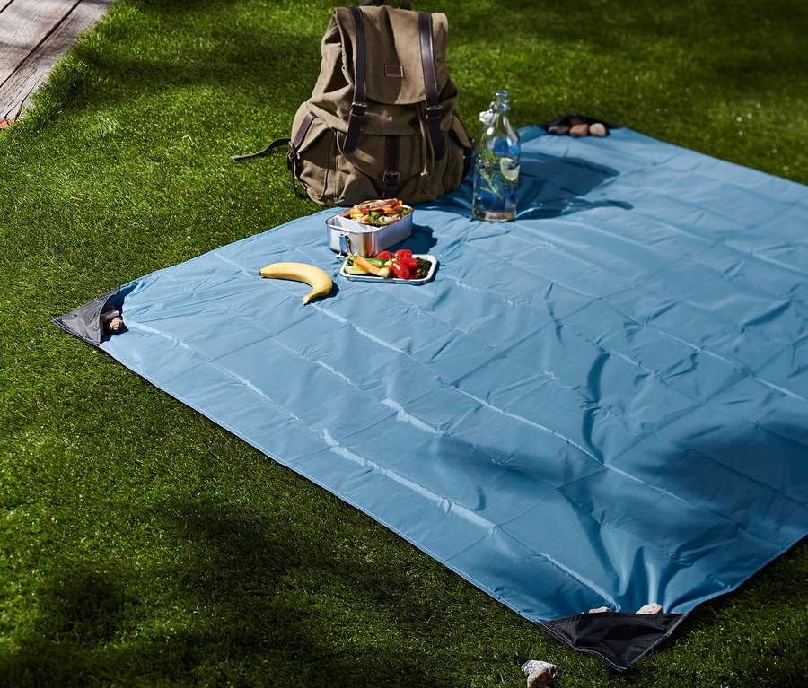 Auf einer Wiese liegt eine blaue Mini-Picknickdecke. Darauf liegen Proviant, eine Banane und eine Wasserflasche. Im Hintergrund steht ein Rucksack.