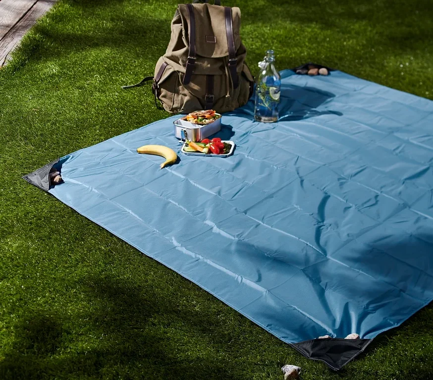 Auf einer Wiese liegt eine blaue Mini-Picknickdecke. Darauf liegen Proviant, eine Banane und eine Wasserflasche. Im Hintergrund steht ein Rucksack.