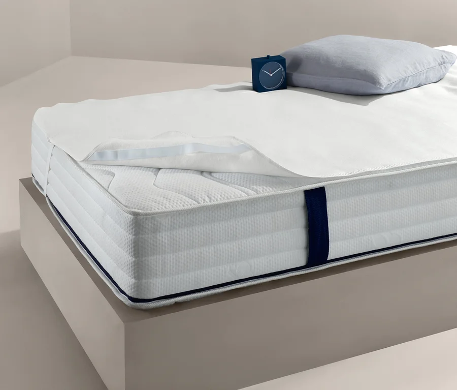 Bett mit weißem Matratzenschoner, ca. 140 x 200 cm, Kissen und Uhr.