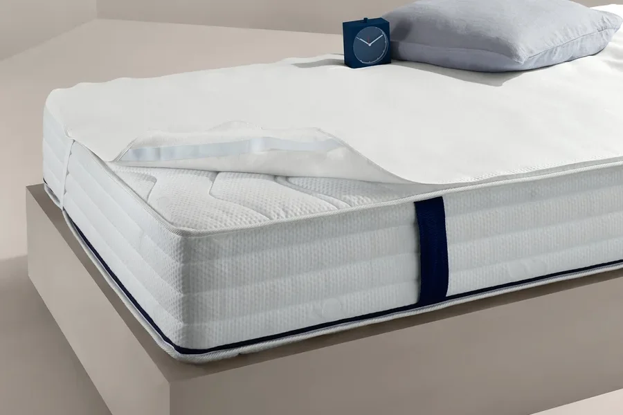 Bett mit weißem Matratzenschoner, ca. 140 x 200 cm, Kissen und Uhr.