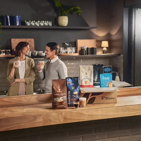 Zwei Personen halten Kaffeetassen in einer Küche hinter Tchibo Kaffeepackungen.