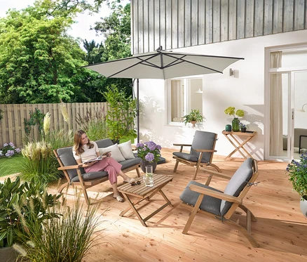 Eine Frau sitzt auf einem zusammenklappbaren Garten-Lounge-Set mit Premium-Kissen, liest und ist umgeben von einem Garten-Tabletttisch und 2 Outdoor-Kissen mit Leinen-Haptik.