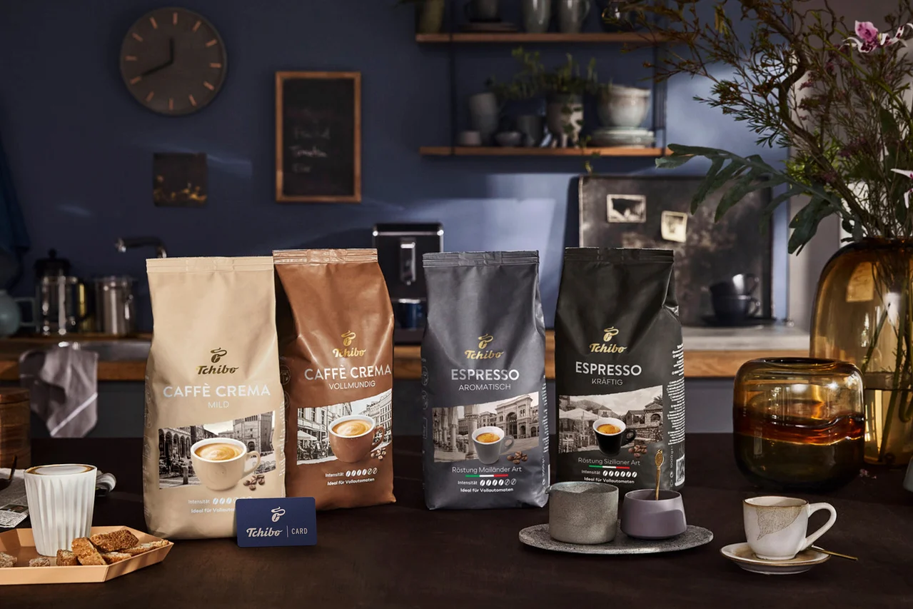 Vier Tchibo Kaffeepackungen, darunter Caffè Crema Mild, Caffè Crema Vollmundig, Espresso Aromtisch und Espresso Kräftig, sind auf einem Tisch ausgestellt. Eine blaue Tchibo Card, eine Kaffeetasse und Biscotti sind ebenfalls vorhanden.