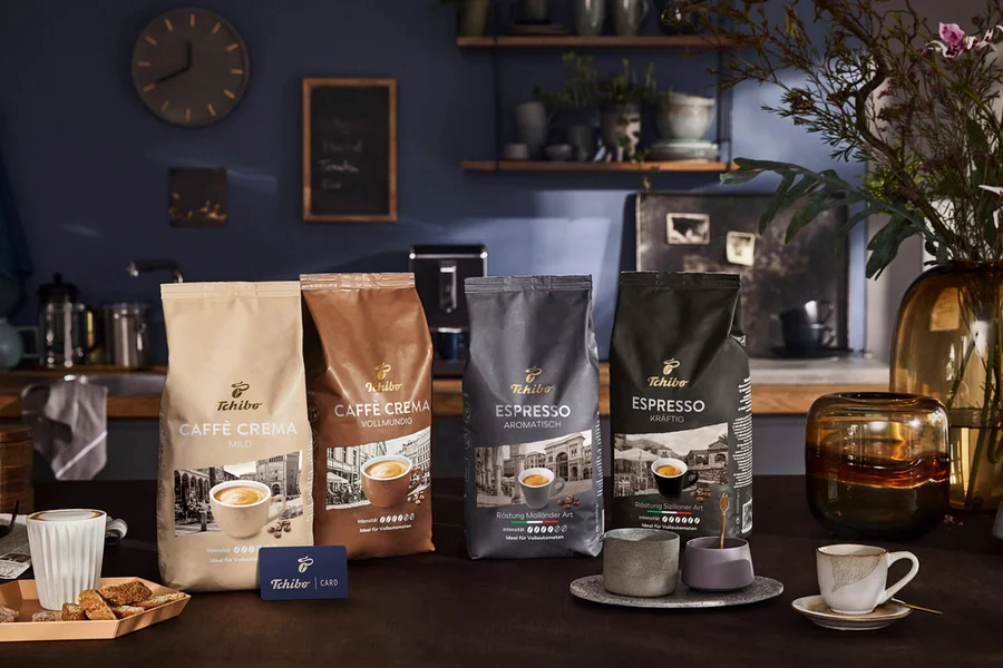 Vier Tchibo Kaffeepackungen, darunter Caffè Crema Mild, Caffè Crema Vollmundig, Espresso Aromtisch und Espresso Kräftig, sind auf einem Tisch ausgestellt. Eine blaue Tchibo Card, eine Kaffeetasse und Biscotti sind ebenfalls vorhanden.