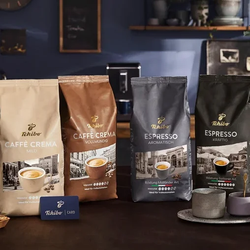 Vier Tchibo Kaffeepackungen, darunter Caffè Crema Mild, Caffè Crema Vollmundig, Espresso Aromtisch und Espresso Kräftig, sind auf einem Tisch ausgestellt. Eine blaue Tchibo Card, eine Kaffeetasse und Biscotti sind ebenfalls vorhanden.