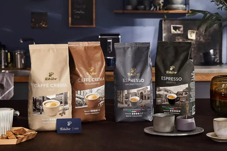 Vier Tchibo Kaffeepackungen, darunter Caffè Crema Mild, Caffè Crema Vollmundig, Espresso Aromtisch und Espresso Kräftig, sind auf einem Tisch ausgestellt. Eine blaue Tchibo Card, eine Kaffeetasse und Biscotti sind ebenfalls vorhanden.