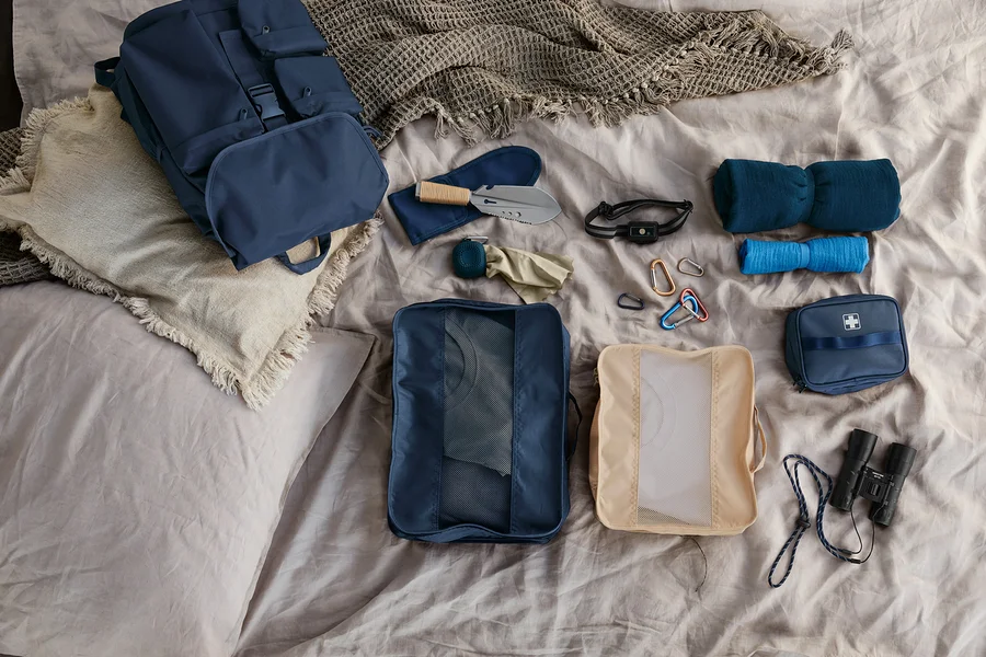 Auf einem Bett liegen ein Outdoor-Rucksack, 5 Karabinerhaken, 2 Musselin-Reisehandtücher, eine Medikamenten-Tasche, ein Outdoor-Handtuch, eine kompakte Stirnlampe, eine 9-in-1-Camping-Schaufel, ein Taschenfernglas »12x32« und 2 Reiseordnungstaschen.