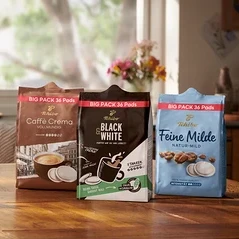 Auf einem Holztisch stehen eine Tasse, ein Teller mit Schokoladenkuchen und drei Packungen Tchibo Kaffeepads: Caffè Crema, Black & White und Feine Milde.