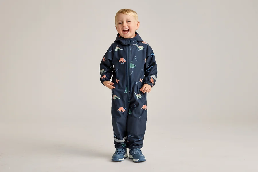 Ein lachender Junge steht in einem marineblauen Overall mit Dinosaurier-Muster und blauen Schuhen vor einem hellen Hintergrund.