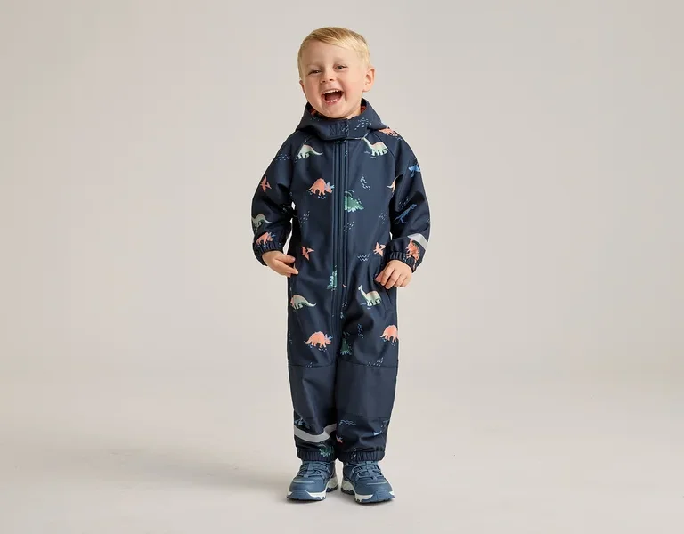 Ein lachender Junge steht in einem marineblauen Overall mit Dinosaurier-Muster und blauen Schuhen vor einem hellen Hintergrund.