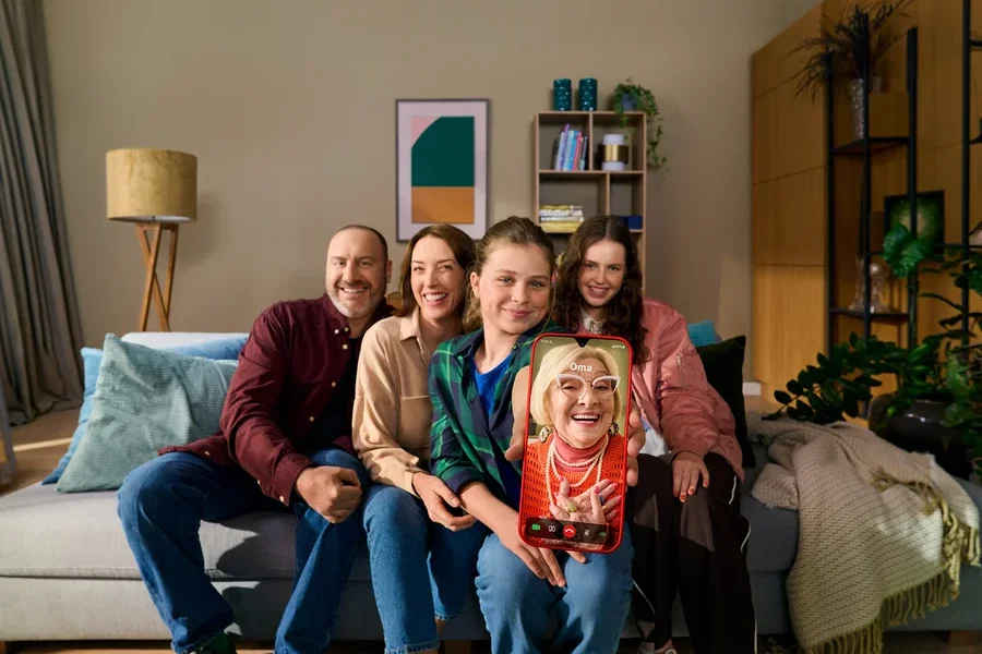 Eine vierköpfige Familie, zwei Erwachsene und zwei Teenager, sitzt lächelnd auf einem Sofa im Wohnzimmer. Ein Teenager hält ein rotes Smartphone mit einem Videoanruf einer älteren Frau, die als „Oma“ beschriftet ist.