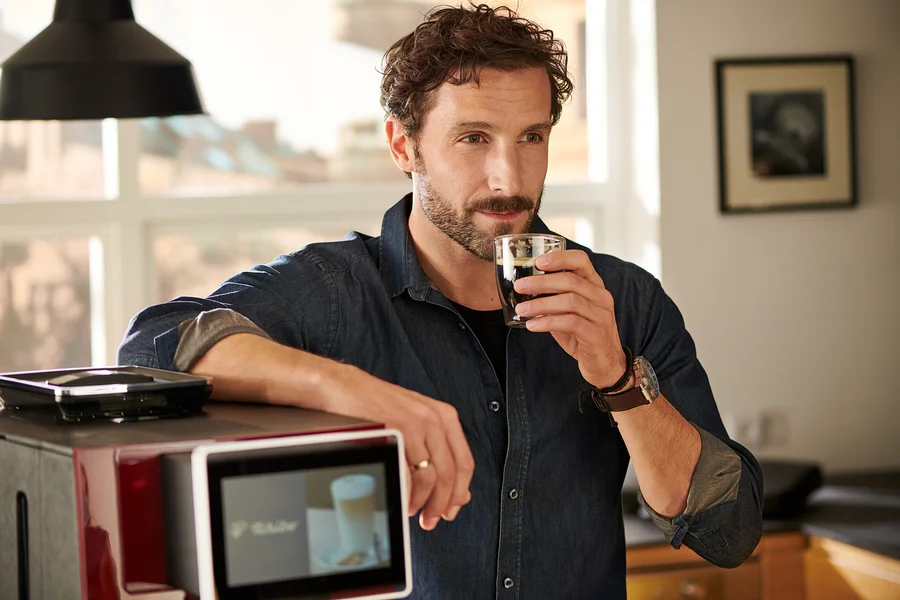 Mann trinkt Kaffee, lehnt an rotem »Tchibo Office« Kaffeevollautomat.