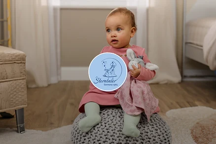 Ein Baby in einem rosa Kleid und hellgrünen Strumpfhosen sitzt auf einem grauen Pouf und hält ein graues Hasen-Stofftier mit einer rosa Decke. Im Hintergrund ist ein Zimmer mit Holzboden und einem Fenster zu sehen.