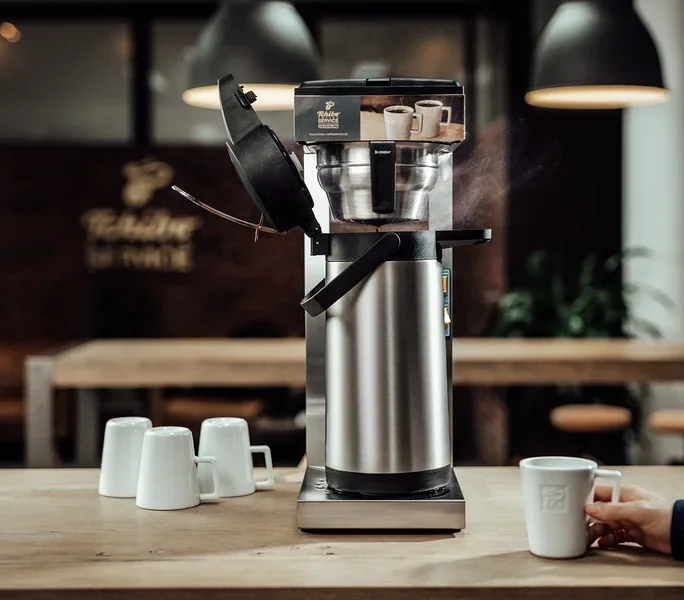 Mann hält Tasse neben »Crem Thermo Tchibo Businessline« Spar-Set Kaffeemaschine. Drei weitere Tassen stehen auf dem Tisch.