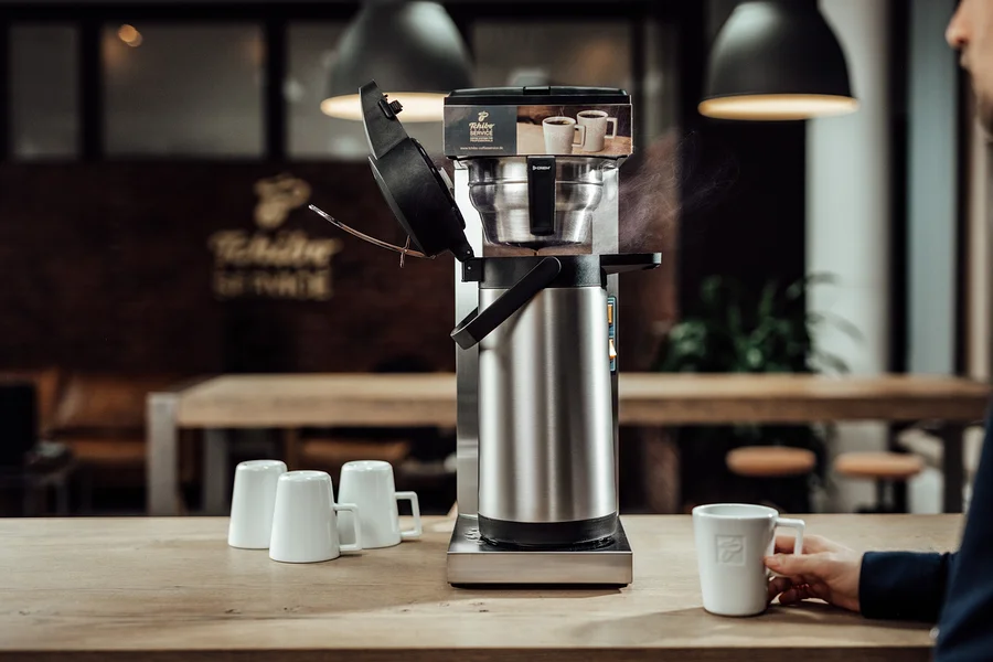 Mann hält Tasse neben »Crem Thermo Tchibo Businessline« Spar-Set Kaffeemaschine. Drei weitere Tassen stehen auf dem Tisch.