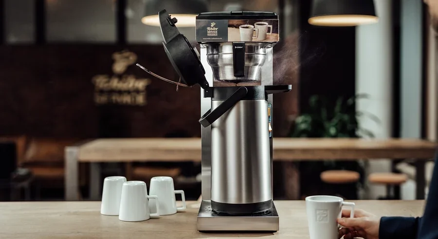 Mann hält Tasse neben »Crem Thermo Tchibo Businessline« Spar-Set Kaffeemaschine. Drei weitere Tassen stehen auf dem Tisch.