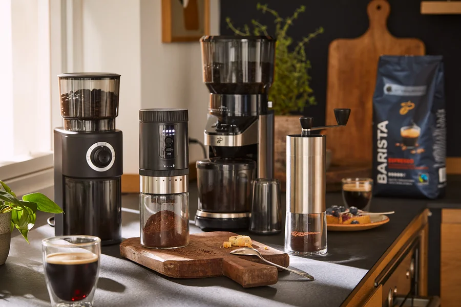 Vier Kaffeemühlen und eine Packung Kaffeebohnen sind auf einer Küchentheke mit Kaffee und Gebäck arrangiert.