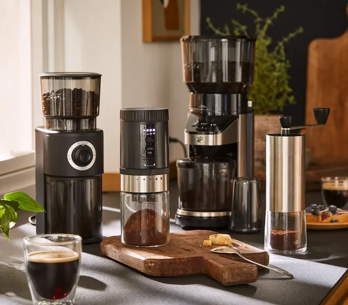 Vier Kaffeemühlen und eine Packung Kaffeebohnen sind auf einer Küchentheke mit Kaffee und Gebäck arrangiert.