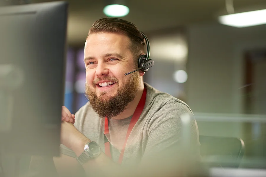 Bärtiger Mann mit Headset lächelt, während er auf einen Computermonitor schaut.