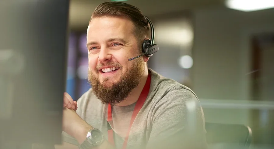 Bärtiger Mann mit Headset lächelt, während er auf einen Computermonitor schaut.
