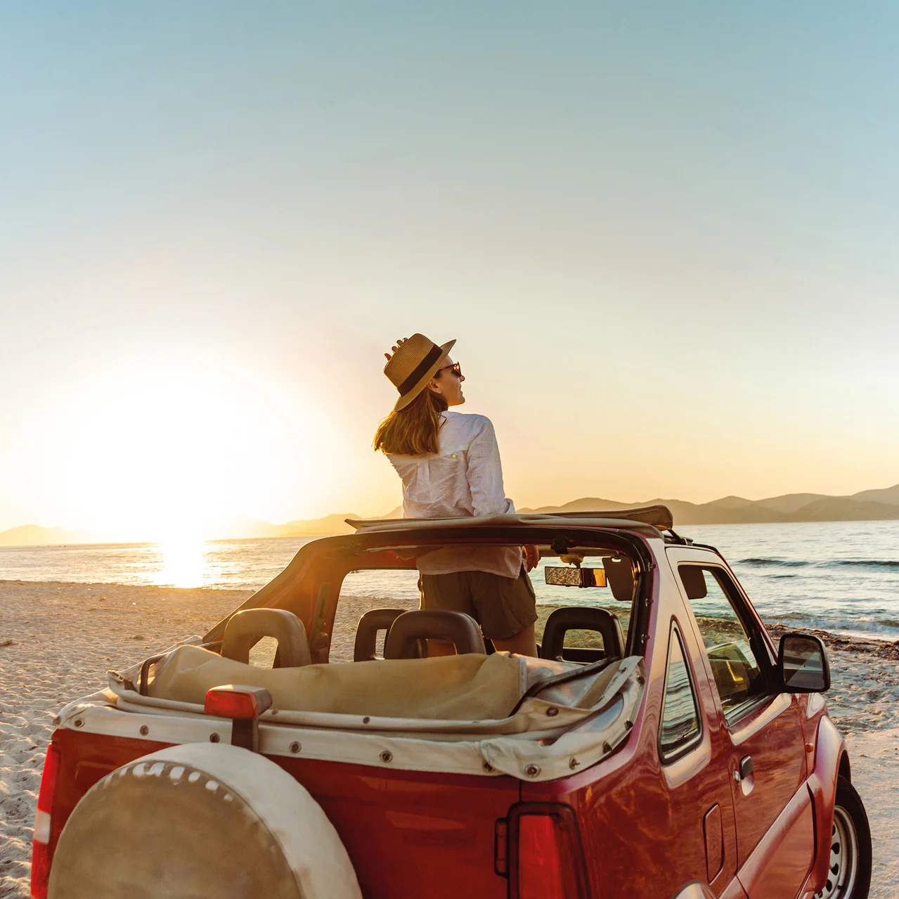 Frau mit Hut und Sonnenbrille steht in rotem Cabrio am Strand und schaut aufs Meer bei Sonnenuntergang.