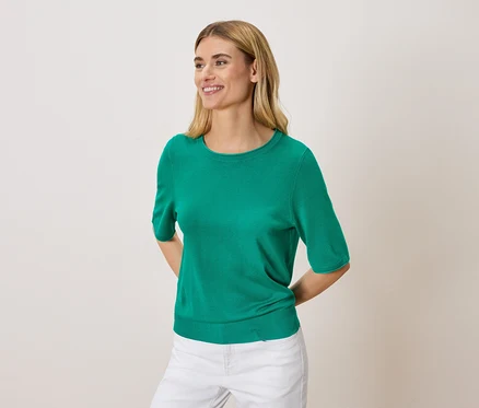 Frau mit blonden Haaren, die einen grünen Pullover und weiße Hosen trägt.