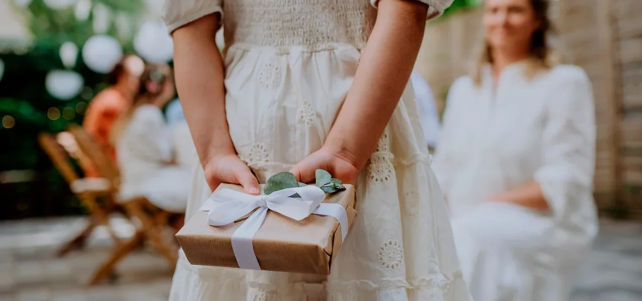 Kind in weißem Lochstickerei-Kleid hält ein braunes Geschenk mit weißer Schleife und Eukalyptusblättern hinter dem Rücken, mit unscharfen Personen im Hintergrund.