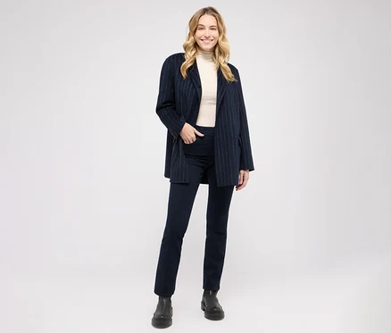 Eine lächelnde blonde Frau steht in einem dunkelblauen Nadelstreifenblazer, einem cremefarbenen Rollkragenpullover, dunklen Jeans und schwarzen Stiefeln.