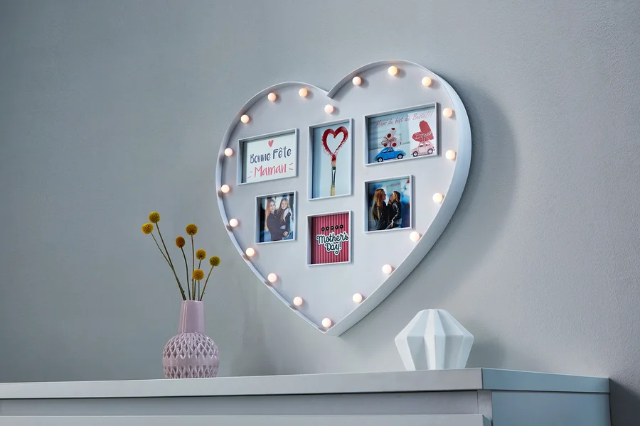 An der Wand hängt ein Herz mit Fotos, das von Glühbirnen beleuchtet wird, darunter eine Vase mit Blumen und ein weißes Dekorationsobjekt.