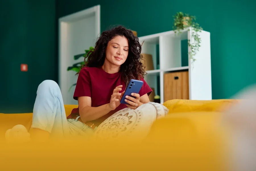 Frau mit lockigem Haar sitzt auf einem gelben Sofa und benutzt ein blaues Smartphone. Im Hintergrund ein Regal und eine grüne Wand.